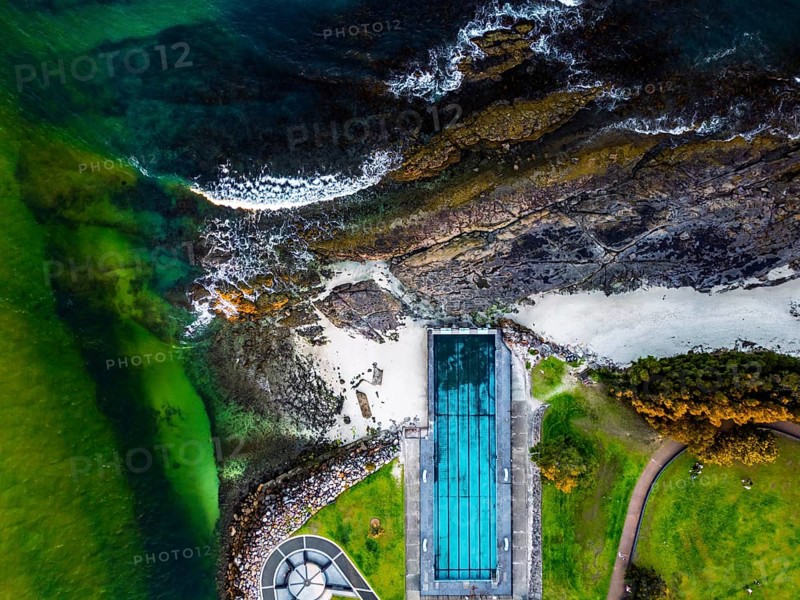 Aerial view capturing the contrast between a coastal pool and rugged shoreline in Jervis Bay, Australia. Vivid greens and blues highlight the natural landscape, offering a striking perspective