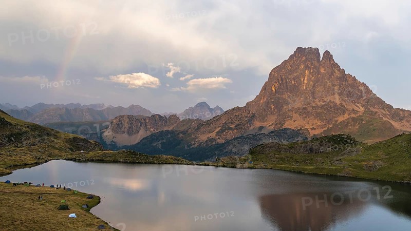 Panoramic view of the Midi d'Ossau peak reflected in a calm lake, surrounded by lush vegetation in the French Pyrenees A serene landscape perfect for nature lovers, with a camping area visible