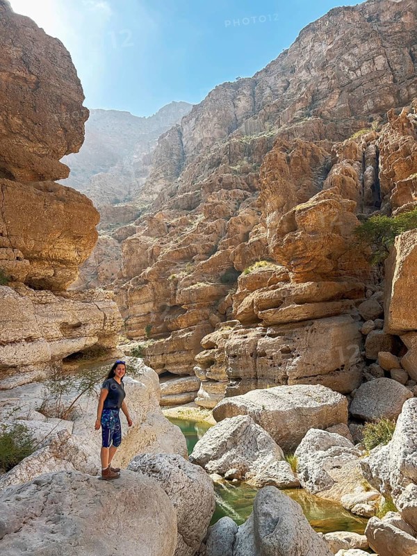 A woman enjoys the scenic beauty of Wadi Ash Shab in Oman, navigating the rocky terrain surrounded by cliffs and clear oasis water pools