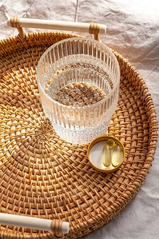 From above a clear glass of water and supplements including Omega 3 and vitamin D3 capsules placed on a rattan tray, embodying a holistic health concept