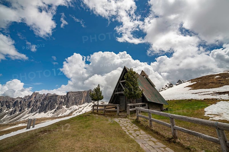 A rustic A-frame hut with a green roof stands on a grassy hill at Passo Giau, surrounded by snow patches and dramatic Dolomite peaks under a dynamic cloudy sky
