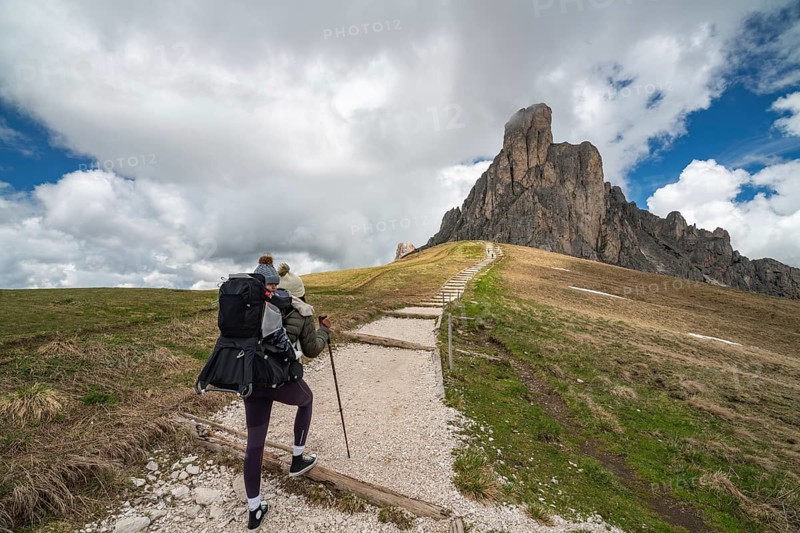A mother with her baby in a carrier hikes along a gravel trail leading to the dramatic rock formations of Passo Giau in the Dolomites, Italy. A beautiful landscape of peaks, sky, and wide-open spaces