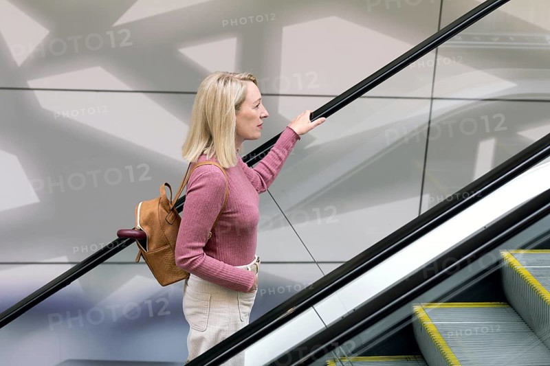Woman rides an escalator in a supermarket, hand on railing, wearing a pink sweater and a brown backpack. Modern architecture and geometric shadows create a dynamic background