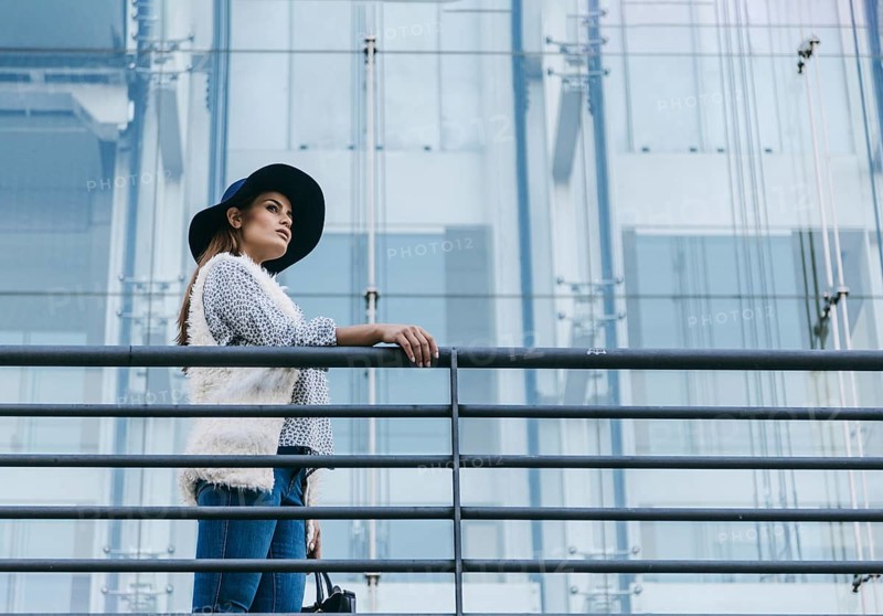 A fashionable woman wearing a stylish hat and chic attire stands confidently next to a modern glass building. Her pose exudes elegance and urban sophistication