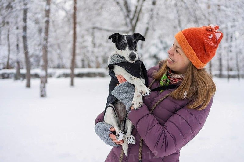 A cheerful woman in a bright hat and warm clothing holds her dog in a snow-covered park. The winter scene reflects warmth and companionship amidst the cold