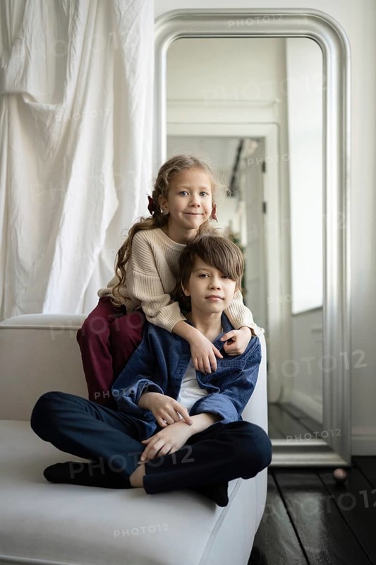 A brother and sister sit together on a white sofa in a bright, minimalistic room The siblings share a close bond, reflecting warmth and happiness in their home environment