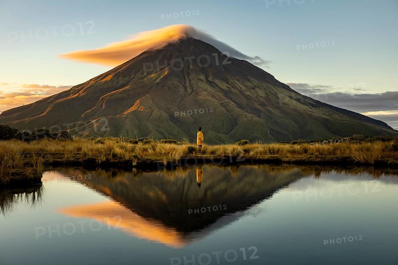 Side view of unrecognizable person standing by a reflective lake, gazing at the majestic Mount Taranaki during sunrise in New Zealand. A serene and tranquil scene unfolds