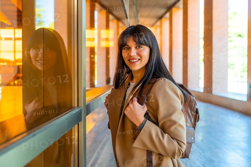 Young woman carrying a backpack and walking along a modern university campus building, smiling confidently while reflecting in the glass, symbolizing education, knowledge, and future success
