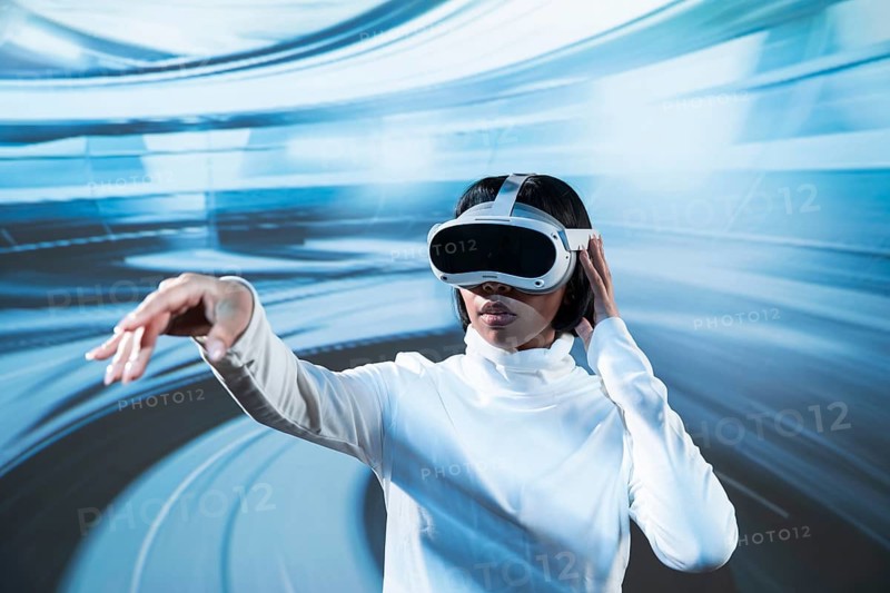 Latin woman in white turtleneck engaging with a virtual environment using VR glasses, reaching out with her hand, set against a dynamic blurred blue motion background