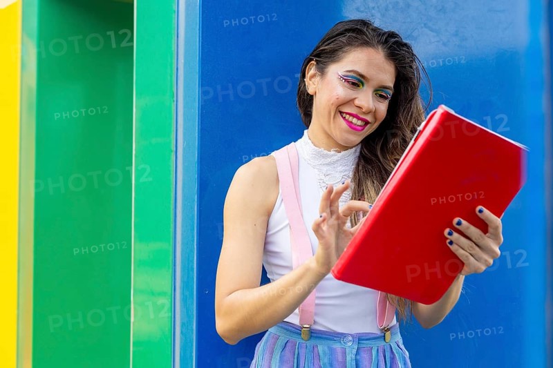 A joyful LGBTQIA+ woman with bold makeup interacts with a red tablet, standing against a vibrant rainbow-colored background. She expresses confidence, creativity, and individuality in a modern setting