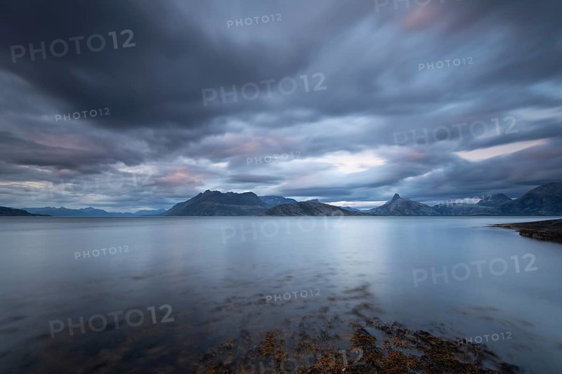 The Vestfjord in Nordland, Norway, offers a dramatic atmosphere near Bodø. Dark clouds are illuminated by the setting sun in shades of red and yellow. The rugged cliffs and mountains are reflected in the quiet fjord, which offers a breathtaking view of Lofoten
