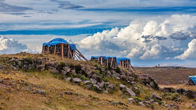 Wooden structures covered by tarps on a hill side under cloudy sky, The Chullpas of Sillustani near Puno in Peru