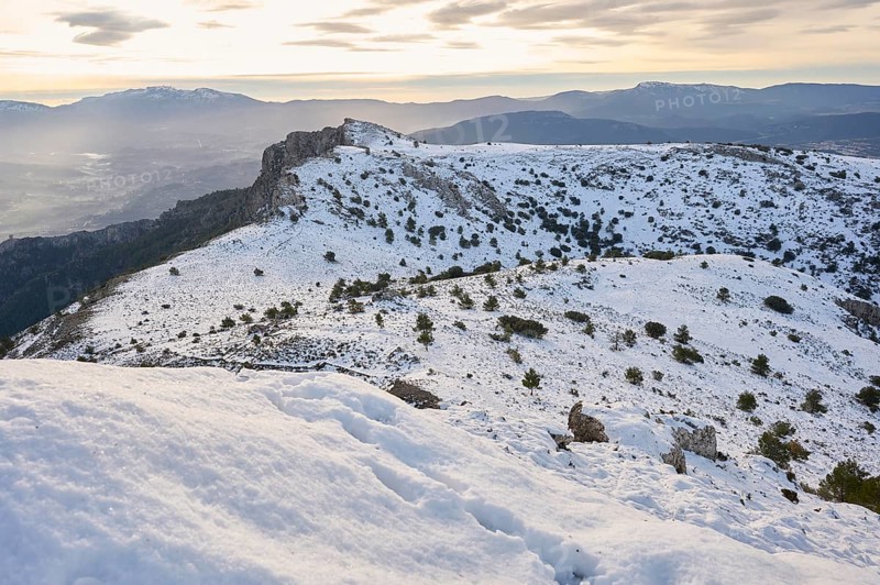 Majestic snowy landscape of Montcabrer, Spain, captures rolling hills covered in snow, dotted with sparse vegetation, under a cloudy sky, offering a tranquil winter view