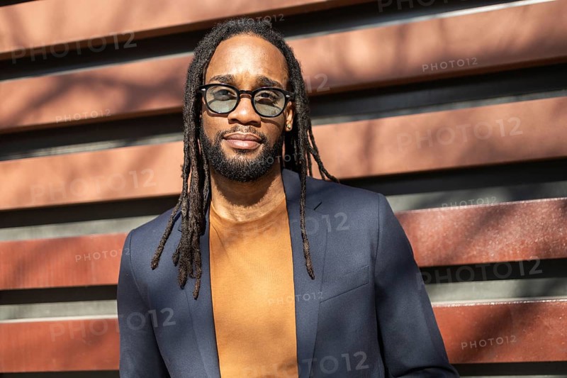 A cuban business man wearing sunglasses and a stylish suit stands against a modern urban backdrop. The sun highlights his expression, embodying professionalism and style