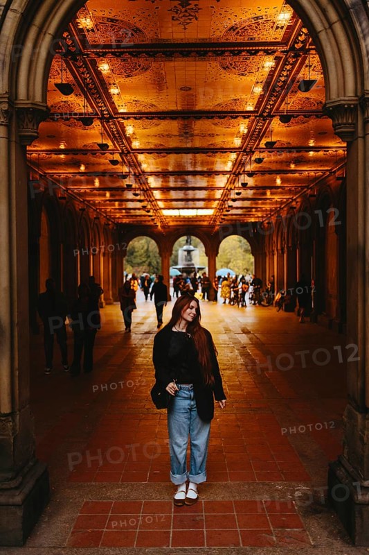 A woman smiles while standing under a illuminated arched corridor with intricate designs in New York. People and a fountain are visible in the background, adding depth to the scene in Central Park