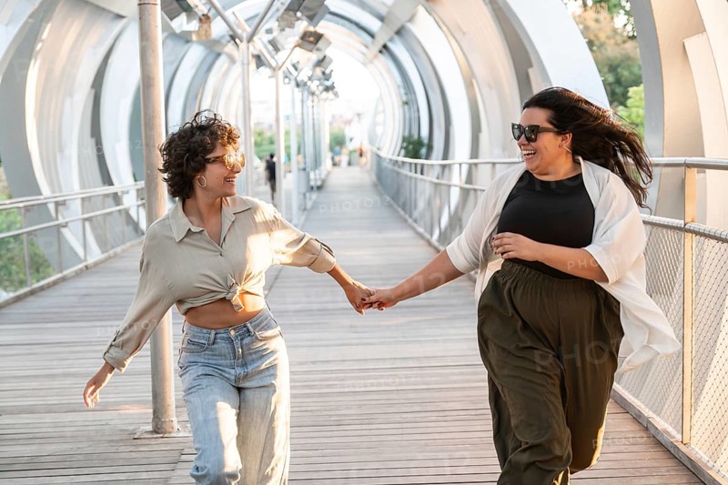 A joyful lesbian couple holding hands and running on a modern urban bridge, radiating happiness and connection. The sun casts a warm glow on their smiling faces