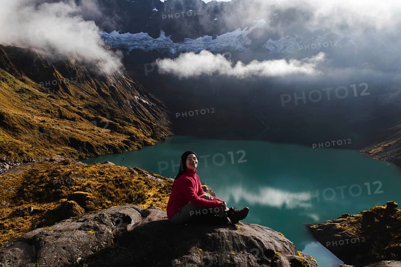 Ecuadorian woman in a red sweater sits on a rock facing a tranquil turquoise lake and lush mountains. Mist hovers over El Altar volcano, Ecuador