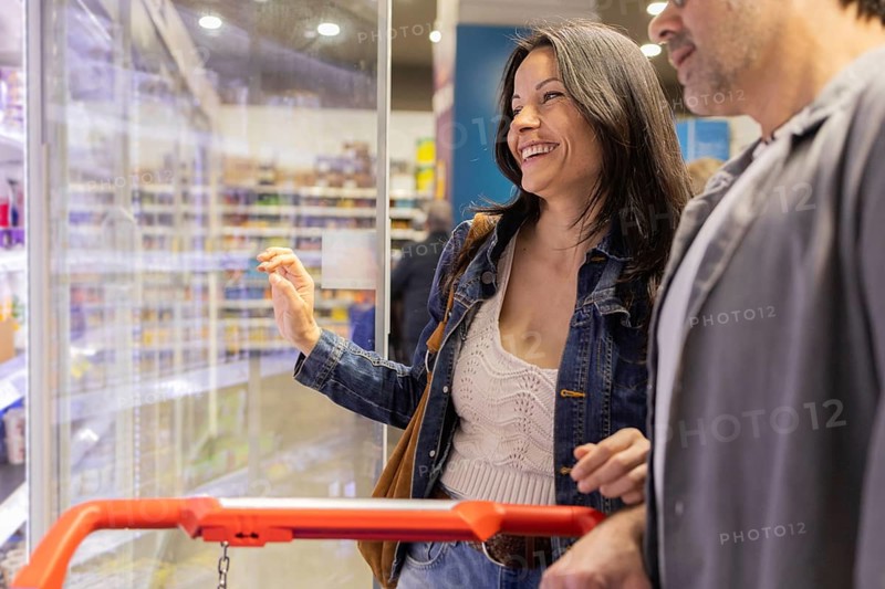 A cheerful couple shops for groceries together in a brightly lit supermarket aisle. They appear engaged and happy as they select items from the shelves, enjoying the experience