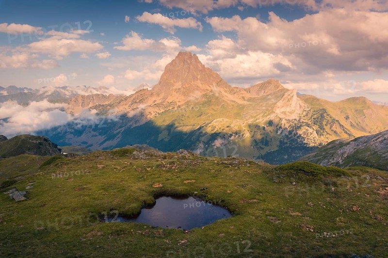 A breathtaking view of a towering mountain under a sky filled with fluffy clouds in summer. A small serene pond sits in the grassy foreground, enhancing the tranquil setting in the Pyrenees