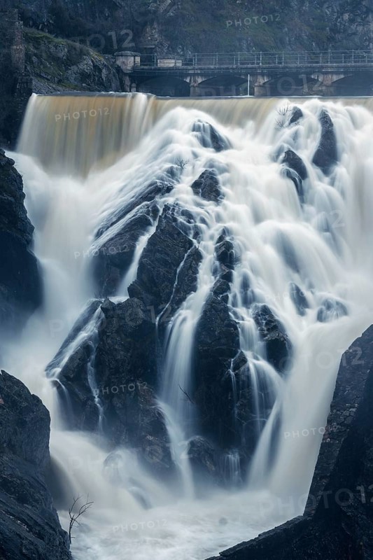 Captivating long exposure photograph showcasing the dynamic waterfall and rocky terrain at Presa o Embalse del Villar, Madrid, under a cloudy sky