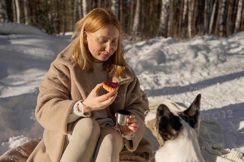 A woman and her Border Collie dog enjoy a sunny winter picnic in the forest. She shares a moment with her pet, feeding him while seated on a blanket covered by snow