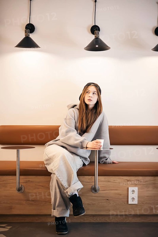 A teenage girl in casual clothes sits thoughtfully on a bench, holding a cup. Her relaxed pose and contemplative expression suggest a quiet moment of reflection