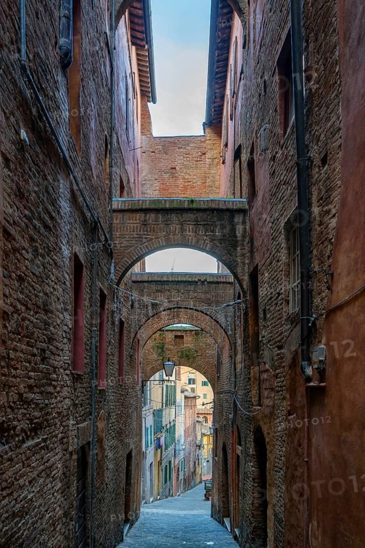 A traditional narrow street in Siena, Tuscany, lined with medieval brick buildings and intricate arches. The alley leads to a colorful view of historic houses bathed in soft light
