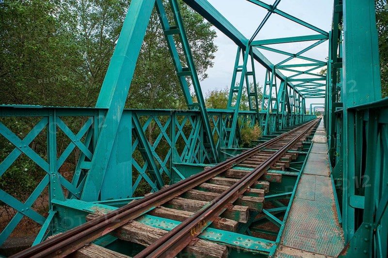 A vibrant green, aged railway bridge spans across a natural landscape, showcasing lattice ironwork and old wooden sleepers amidst lush greenery