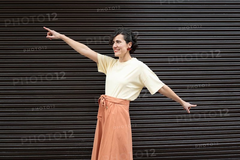 A cheerful woman in a light shirt and orange skirt stands in front of a striped background, energetically pointing with both hands. Her facial expression radiates positivity