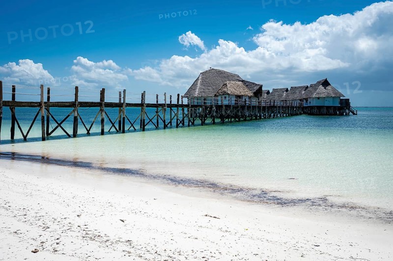 A long wooden pier leads to traditional thatched-roof bungalows built on stilts over the turquoise ocean under a bright blue sky. In Zanzibar, Tanzania