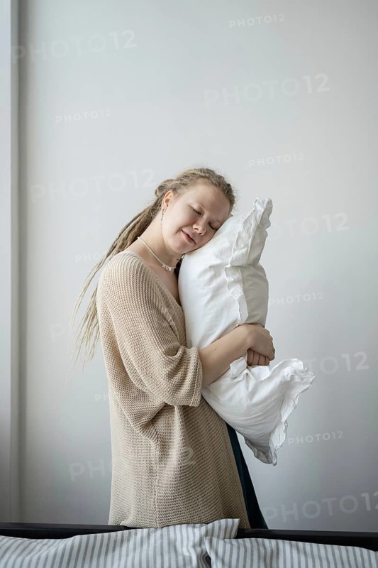 A woman with closed eyes lovingly cuddles a soft white pillow, exuding a sense of peace and contentment She wears a beige sweater, creating a tranquil atmosphere