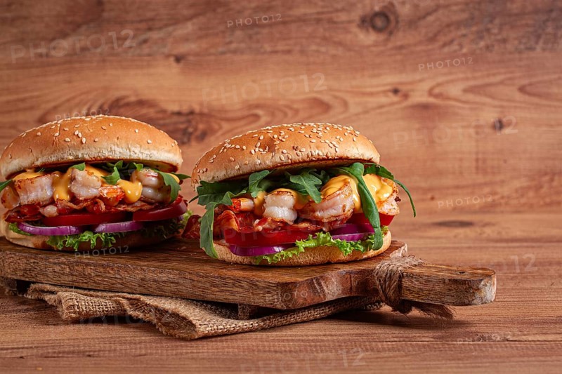 Two shrimp burgers and buns with sesame seeds, bacon, lettuce, tomatoes and onions, on a wooden board, rustic style