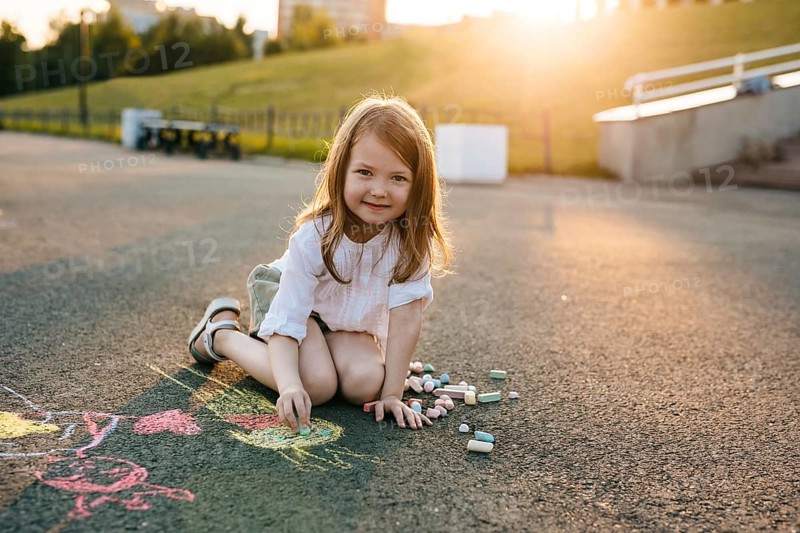 A child sits on pavement, happily drawing with colorful chalk in a sunny park. The scene captures a joyful moment of outdoor creativity and play in summer