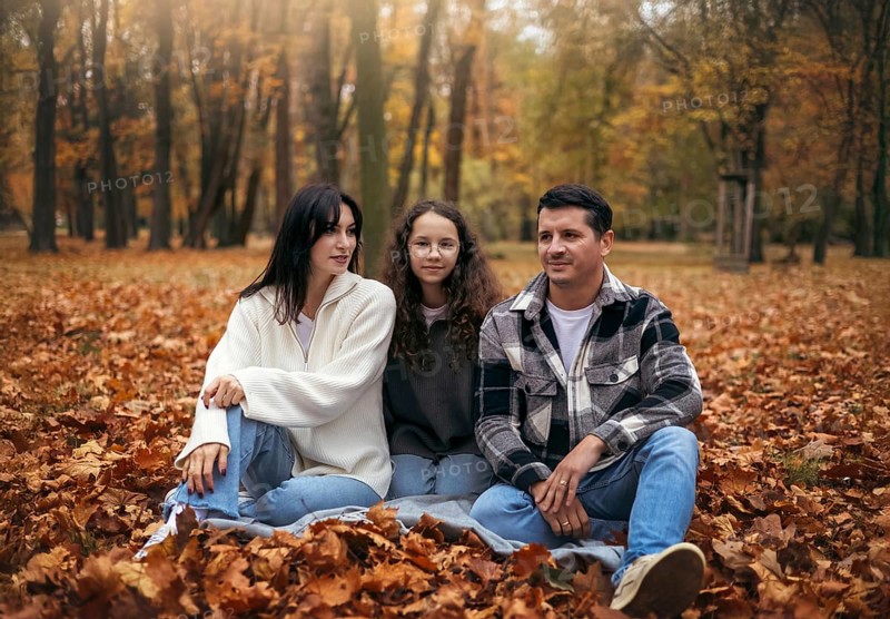 A family sits together among fallen leaves on a crisp autumn day in a tree-filled park. They are warmly dressed, reflecting a cozy and peaceful atmosphere