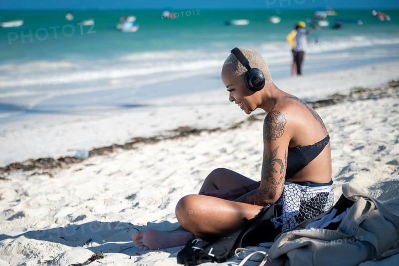PAJE, ZANZIBAR - OCTOBER 18, 2025: A young woman with tattoos sitting on a white sand beach, wearing headphones and enjoying music against a backdrop of turquoise ocean waters