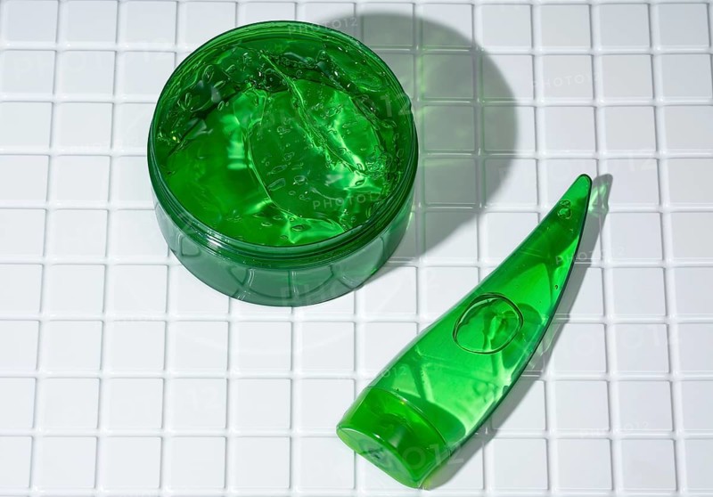 Open container of vibrant green aloe gel next to a body care cream tube on a white tiled bathroom surface, indicating natural skin treatment
