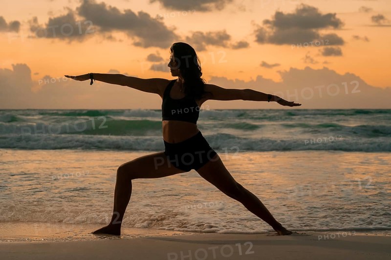 A woman practices yoga in the warrior pose on a sandy beach at sunrise, silhouetted against a colorful sky. The calm ocean waves enhance the serene atmosphere of the scene