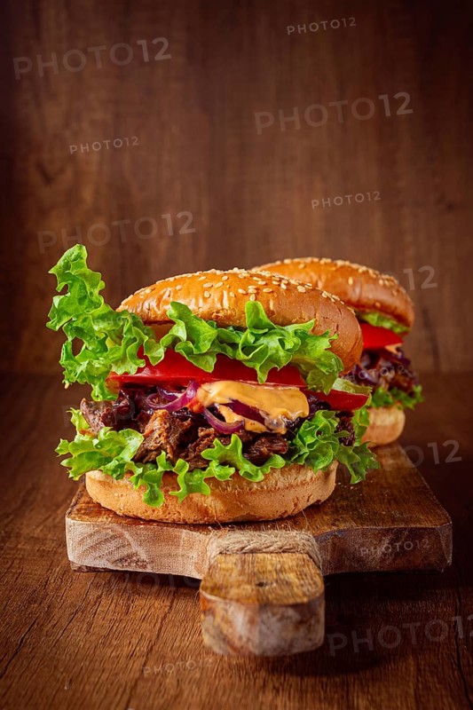 Two burgers are stacked high with fresh lettuce, tomato slices, grilled onions, and a savory sauce. They sit on a wooden board in a warm light setting. Ideal for a meal or snack
