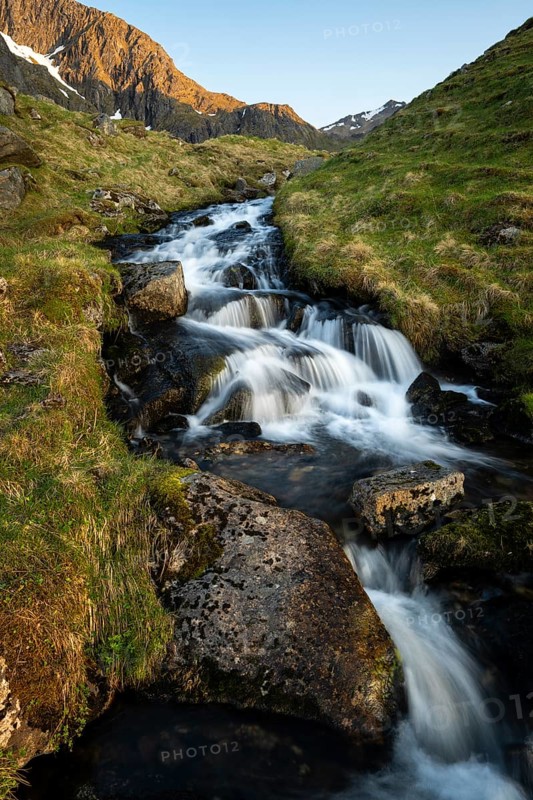 A torrent flows down the mountain. Above left the mountain Stornappstinden. Long exposure. At night at the time of the midnight sun in good weather. Early summer. Flakstadoya, Lofoten, Norway