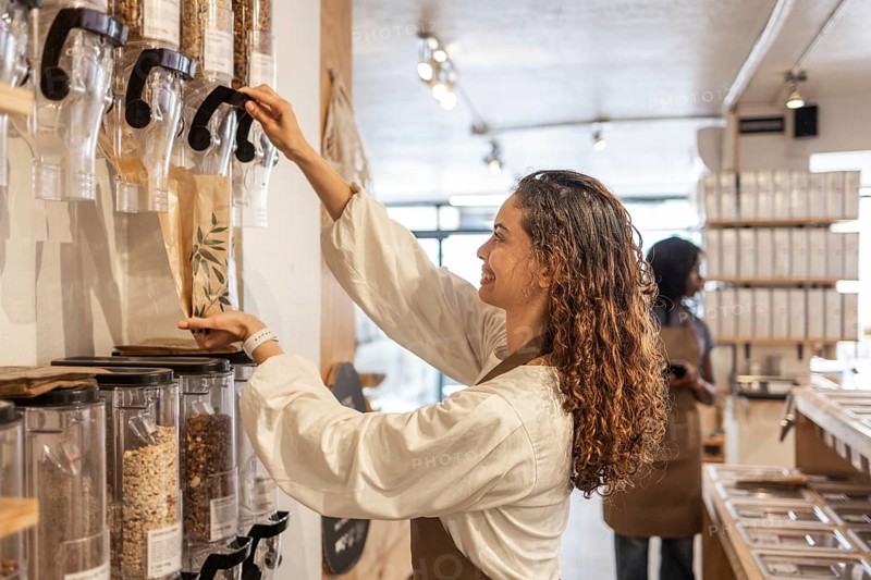 A smiling clerk fills a paper bag with grains from dispensers in an organic supermarket's bulk section. Shelves in the background suggest a well-stocked store