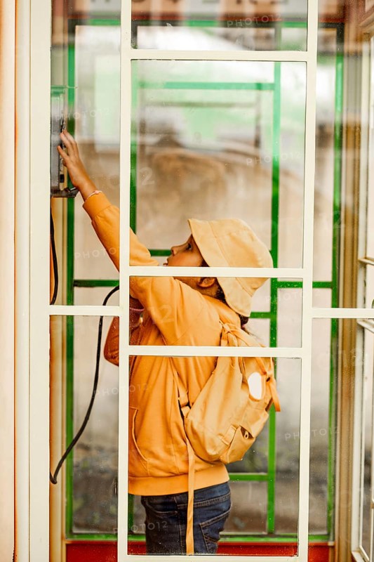 Side view of a preteen girl wearing a yellow hat and hoodie dialing on a vintage phone mounted on a red wall