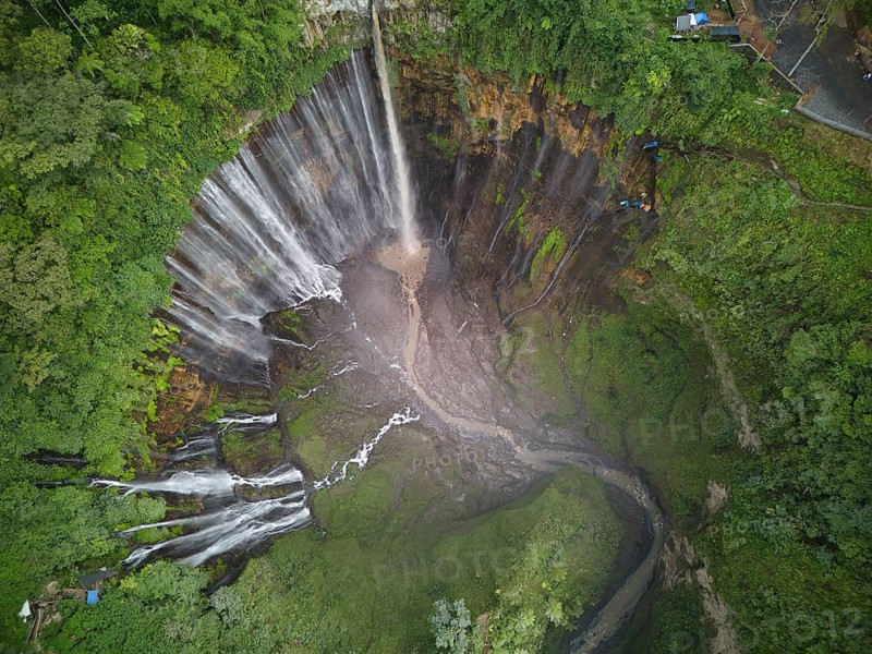 A breathtaking aerial photo capturing the vast Tumpak Sewu Waterfall surrounded by dense tropical forest. The cascade falls powerfully into a natural amphitheater of jagged cliffs and lush vegetation