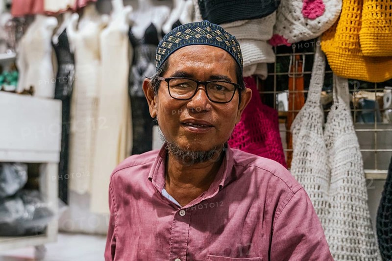 A balinese man in a pink shirt smiles while standing in front of colorful crocheted bags at a market stall, showcasing diverse handmade crafts
