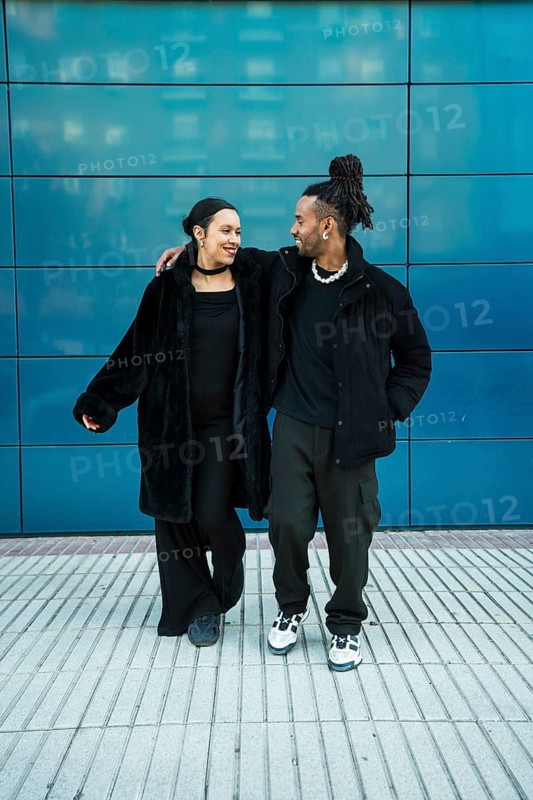 A multicultural couple embraces, showcasing their connection and diversity. The man wears casual streetwear, and the woman dons a chic coat against an urban backdrop