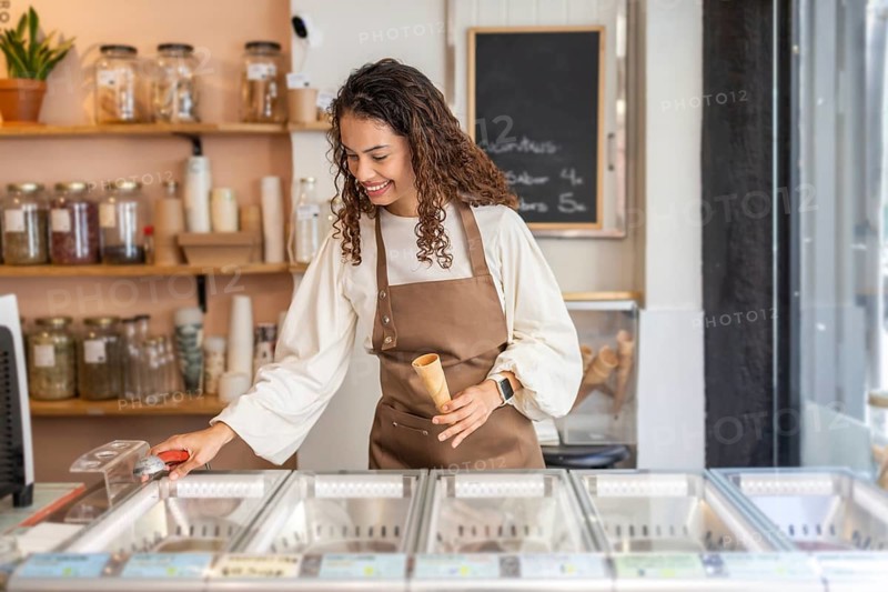 A friendly clerk in an apron serves ice cream in a cozy organic supermarket. She holds a cone while preparing a scoop, surrounded by bulk jars on wooden shelves