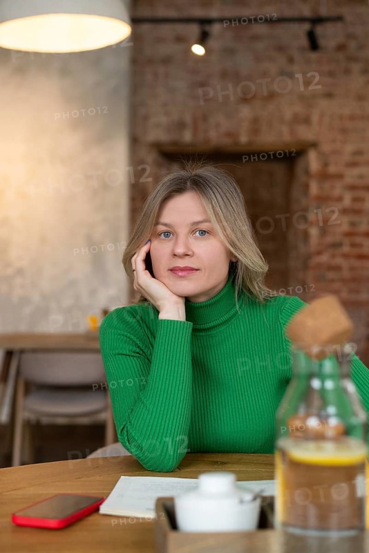 A woman in a green sweater sits at a cafe table, surrounded by a warm, cozy ambiance. With her hand resting on her face, she appears relaxed and thoughtful