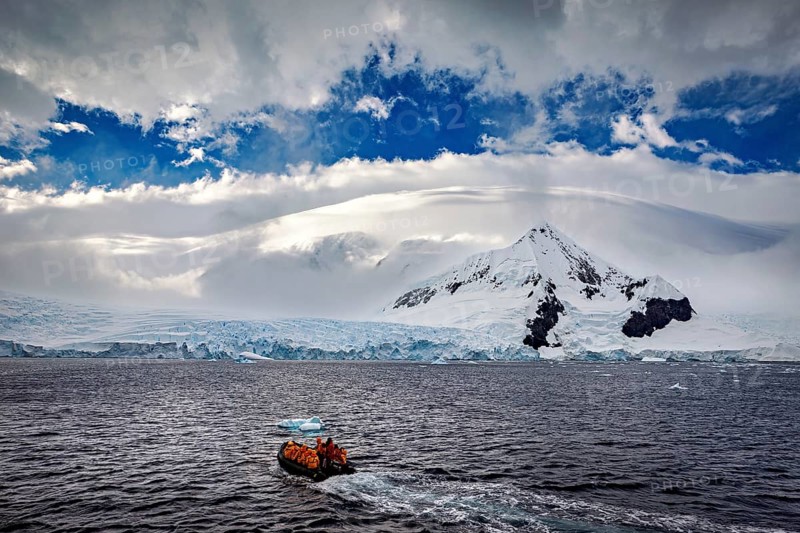 A small boat sails across the sea against a dramatic backdrop of snow-covered mountains and clouds, people in a rubber dinghy travelling on the sea in the Southern Ocean in Antarctica