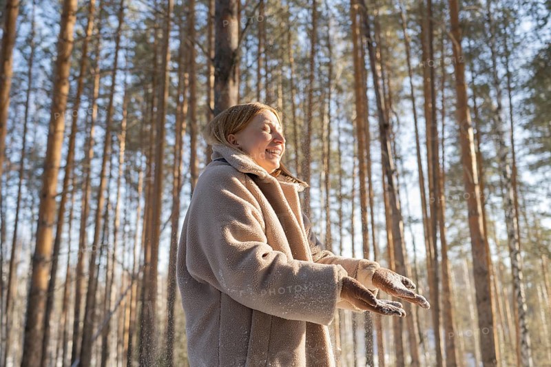 A woman smiles joyfully as she experiences the winter beauty in a sunlit forest. The warmth of her coat contrasts with the snowy setting