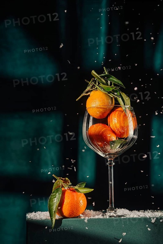 Fresh tangerines with leaves in a tall wine glass, set against a dark background with shards of ice glistening around, evoking a cozy winter atmosphere