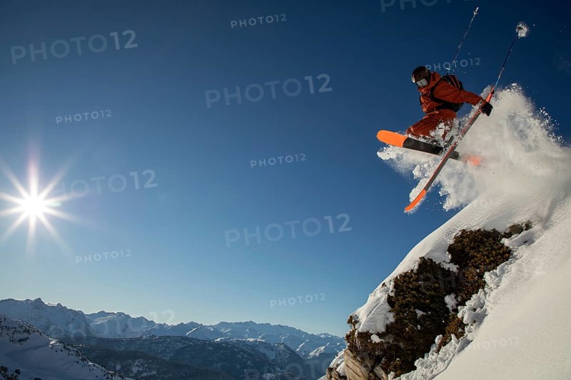 A skier wearing bright gear takes an exhilarating jump off a snowy cliff, surrounded by majestic mountains and a clear blue sky The sun shines brightly overhead
